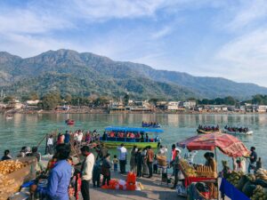 Crowd at a riverside ghat in Rishikesh with food stalls, boats, and people enjoying rafting on the Ganges river against a backdrop of forested hills.