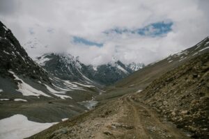 Snow-covered rocky mountains and a remote dirt road winding through a high-altitude valley in Ladakh under cloudy skies.