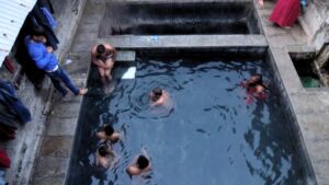 People enjoying the Vashisht Hot Springs, with several individuals swimming in a stone-lined hot water pool and others sitting on the edges, surrounded by a rustic setting.