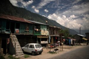  A rustic street scene in Old Manali with wooden houses, a parked white car, clothes hanging outside, and a person standing on a roof, with mountains in the background under a partly cloudy sky.