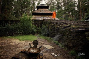 A person in an orange robe standing on a stone structure near a temple with a wooden roof, surrounded by dense forest, with incense burning in the foreground.