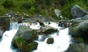 A scenic view of Jogini Waterfalls with water cascading over rocks into a stream, surrounded by lush green vegetation and moss-covered stones.