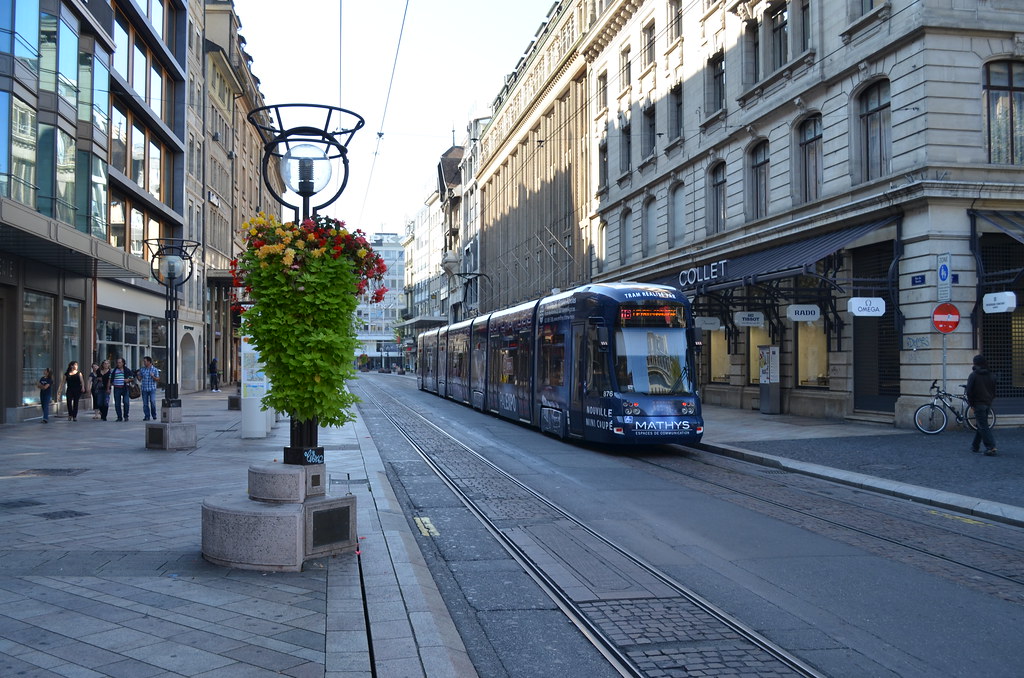 A vibrant street view in Geneva, Switzerland, featuring a modern tram traveling along a city road lined with elegant buildings. A flower-decorated street lamp stands prominently, with pedestrians walking on the sidewalk and a cyclist nearby, all set against a backdrop of classic architecture.