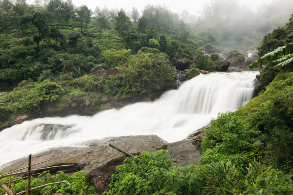A scenic view of Attukad Waterfalls in Munnar, India, featuring a cascading waterfall surrounded by lush green vegetation and rocky terrain. The misty hills and dense forest in the background enhance the natural beauty, with a clear stream flowing over rocks, creating a tranquil atmosphere under a foggy sky.