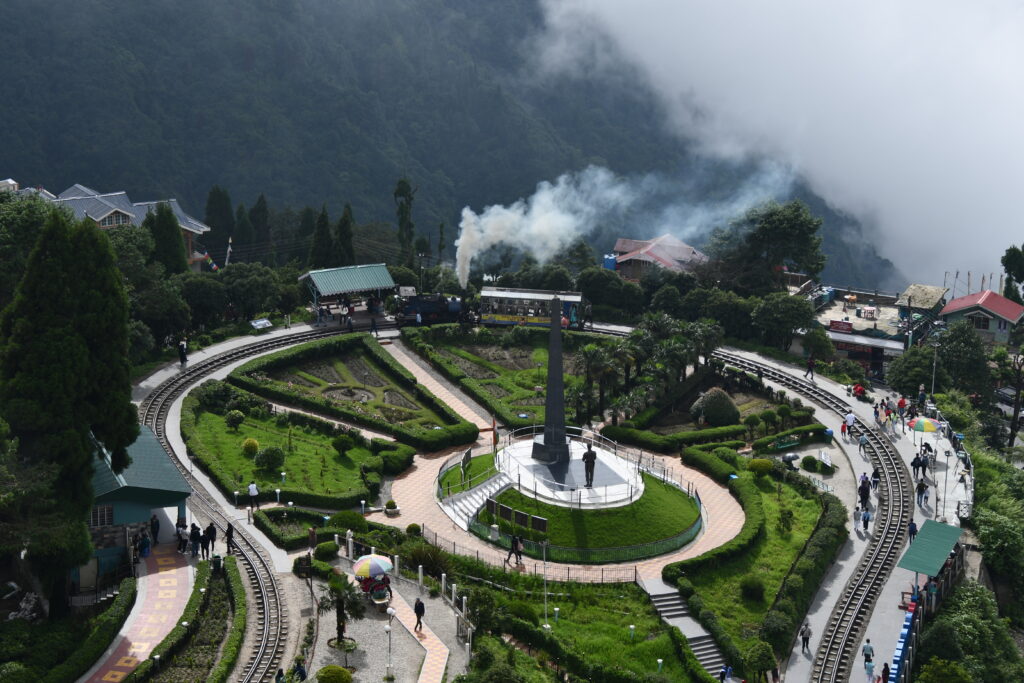 A serene white Peace Pagoda nestled among lush green trees on a hillside at Batasia Loop, Darjeeling, with a small house and clear blue sky in the background.