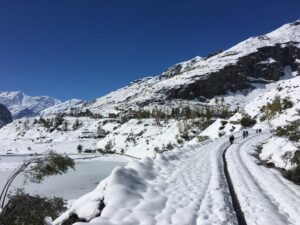 A snowy landscape of Rohtang Pass with a winding snow-covered path, flanked by snow-laden hills and a frozen lake, with a few people walking in the distance under a clear blue sky.