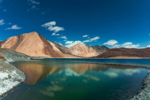 A serene lake in Ladakh reflecting the surrounding barren mountains and clear blue sky with scattered clouds.