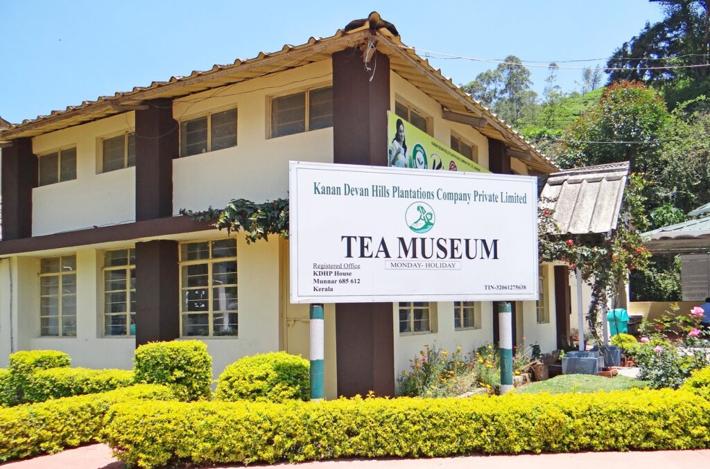 The Munnar Tea Museum building in India, managed by Kanan Devan Hills Plantations Company Private Limited, featuring a signboard with details. The structure has a tiled roof, white walls with brown pillars, and large windows. The foreground showcases well-maintained yellow-green hedges and colorful flower beds, with lush green hills in the background under a clear blue sky.