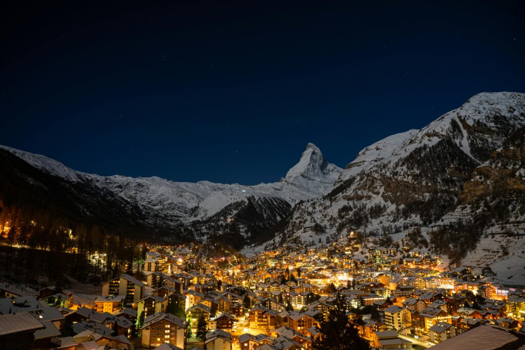A stunning nighttime view of Zermatt, a picturesque town in Switzerland, nestled in a valley surrounded by snow-covered mountains. The town is illuminated with warm lights from numerous buildings, contrasting against the dark, starry sky and the prominent Matterhorn peak in the background.