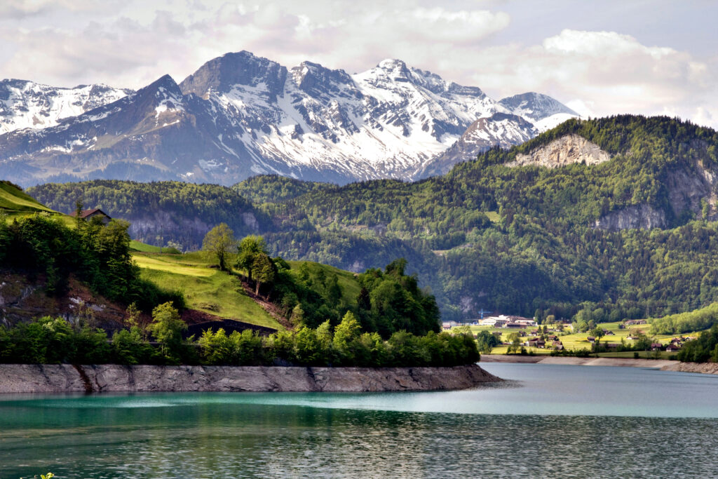 A scenic autumn view of Interlaken, Switzerland, featuring elegant historic buildings with a prominent central structure, surrounded by trees with vibrant fall colors. Snow-capped mountains rise in the background, partially shrouded in mist, under a cloudy sky.