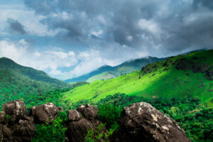 A panoramic view of the lush green hills of Coorg, Karnataka, with rocky outcrops in the foreground and misty mountains under a cloudy sky.