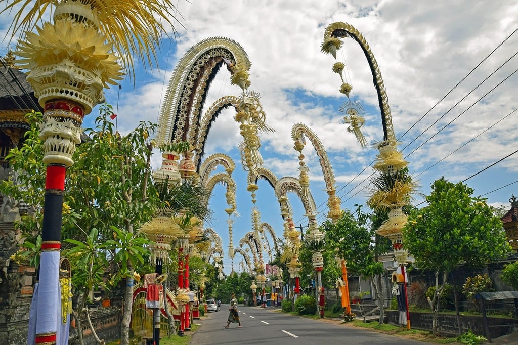 A vibrant street scene in Bali, Indonesia, adorned with traditional Penjor decorations, featuring arched bamboo structures with intricate designs, set against a backdrop of lush greenery and a cloudy sky.
