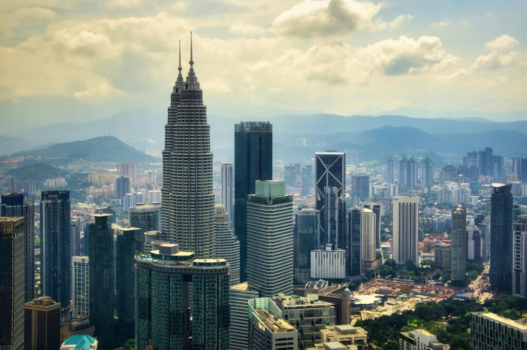 A stunning aerial view of the Kuala Lumpur skyline featuring the iconic Petronas Towers, surrounded by modern skyscrapers and lush green hills under a cloudy sky.