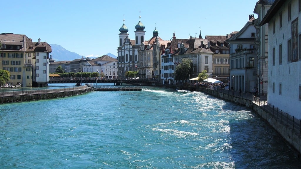 A beautiful daytime view of Lucerne, Switzerland, featuring traditional wooden chalets with sloped roofs nestled in a lush valley. Snow-capped mountains rise prominently in the background under a partly cloudy sky.
