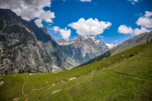  scenic view of a lush green valley with rugged mountains, snow-capped peaks, and a clear blue sky with scattered clouds.