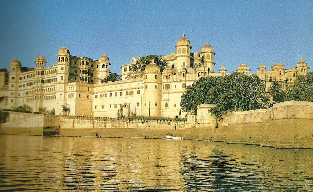A grand view of the City Palace in Udaipur, Rajasthan, India, showcasing a large light-colored palace with multiple domes, towers, and arched windows, situated along the edge of Lake Pichola with calm water in the foreground and a clear blue sky above.