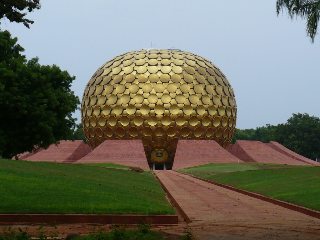 A striking view of the Matrimandir, a golden spherical structure with a pattern of circular discs, located in Auroville, India. The building is set against a backdrop of green lawns and trees, with a red stone pathway leading to its entrance. The sky above is clear with a soft blue hue.