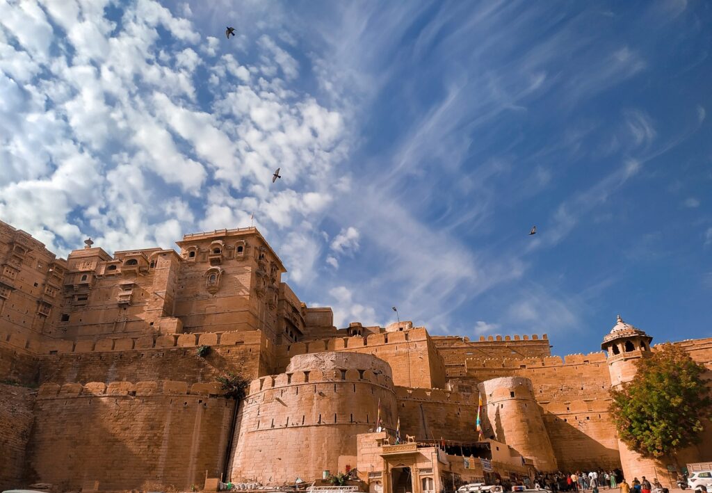 A majestic view of Jaisalmer Fort in Rajasthan, India, a massive golden sandstone structure with towering walls, bastions, and intricate carvings, rising against a bright blue sky with wispy clouds and birds soaring overhead, people and vehicles visible at the base entrance.