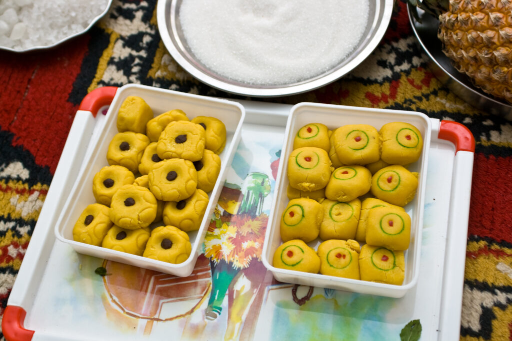 Two trays of milk peda sweets, one with plain yellow pedas and the other with colorful green and red designs, placed on a decorative tray with sugar and a pineapple in the background.
