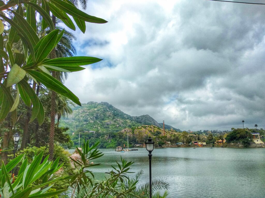 Serene Nakki Lake in Mount Abu, Rajasthan, surrounded by lush greenery, hills, and a cloudy sky with palm trees in the foreground.