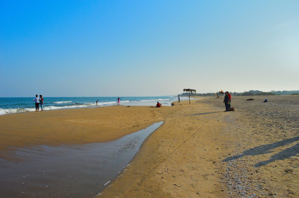A serene view of Paradise Beach in Puducherry, featuring a wide sandy shoreline with gentle waves lapping at the shore. A small stream of water flows across the sand, and a few people are scattered along the beach, some walking and others sitting. The sky is clear and blue, with a thatched umbrella and distant greenery visible in the background.