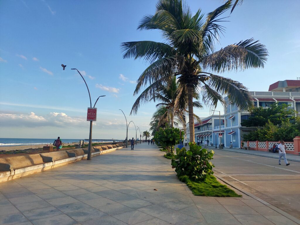 A scenic view of the Puducherry promenade featuring a paved walkway lined with tall palm trees, modern street lamps, and colorful buildings. The pathway runs parallel to a sandy beach with gentle waves, and a few people are seen walking or sitting. The sky is clear with a few clouds, and a bird is flying overhead.