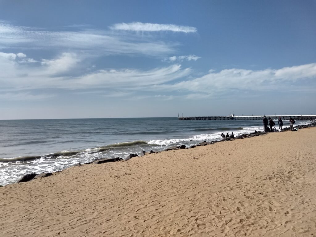 A picturesque view of Promenade Beach in Puducherry, featuring a wide paved walkway lined with tall palm trees, modern street lamps, and colorful buildings. The pathway runs alongside a sandy beach with gentle ocean waves, and a few people are walking or sitting. The sky is clear with scattered clouds, and a bird is flying overhead.