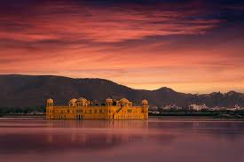 A serene image of Jal Mahal, a stunning palace in Rajasthan, India, situated in the middle of Man Sagar Lake, with its reflection visible in the calm water. The sky is painted with vibrant hues of pink, orange, and purple during sunset, and distant hills form the backdrop.