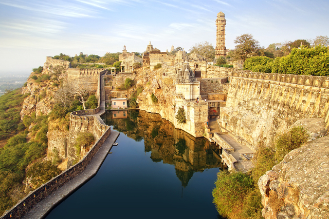 Aerial view of the historic Chittorgarh Fort in Rajasthan, India, perched on a cliffside with ancient stone walls, temples, a tall victory tower, and a reflective water reservoir below, under a clear blue sky.