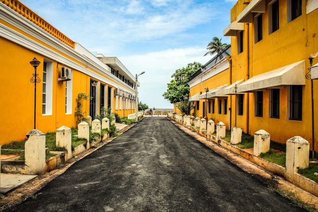 A serene view of Paradise Beach in Puducherry, featuring a wide sandy shoreline with gentle waves lapping at the shore. A small stream of water flows across the sand, and a few people are scattered along the beach, some walking and others sitting. The sky is clear and blue, with a thatched umbrella and distant greenery visible in the background.