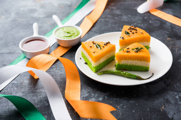 A plate with three pieces of tricolour dhokla, featuring green, white, and orange layers, garnished with spices and a green chili, served with small bowls of chutney, surrounded by tricolour ribbons.