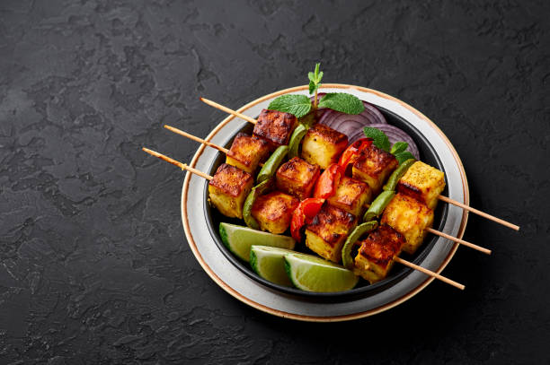 A plate of Tiranga Paneer Tikka skewers with marinated paneer, bell peppers, and onions, garnished with lime wedges and mint, served on a dark textured surface.
