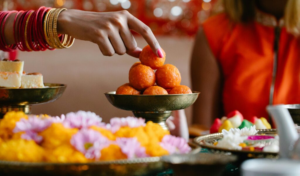 A hand adorned with red and gold bangles reaching for a stack of orange motichur laddus in a decorative metal bowl, surrounded by other Indian sweets and flowers on a festive table.