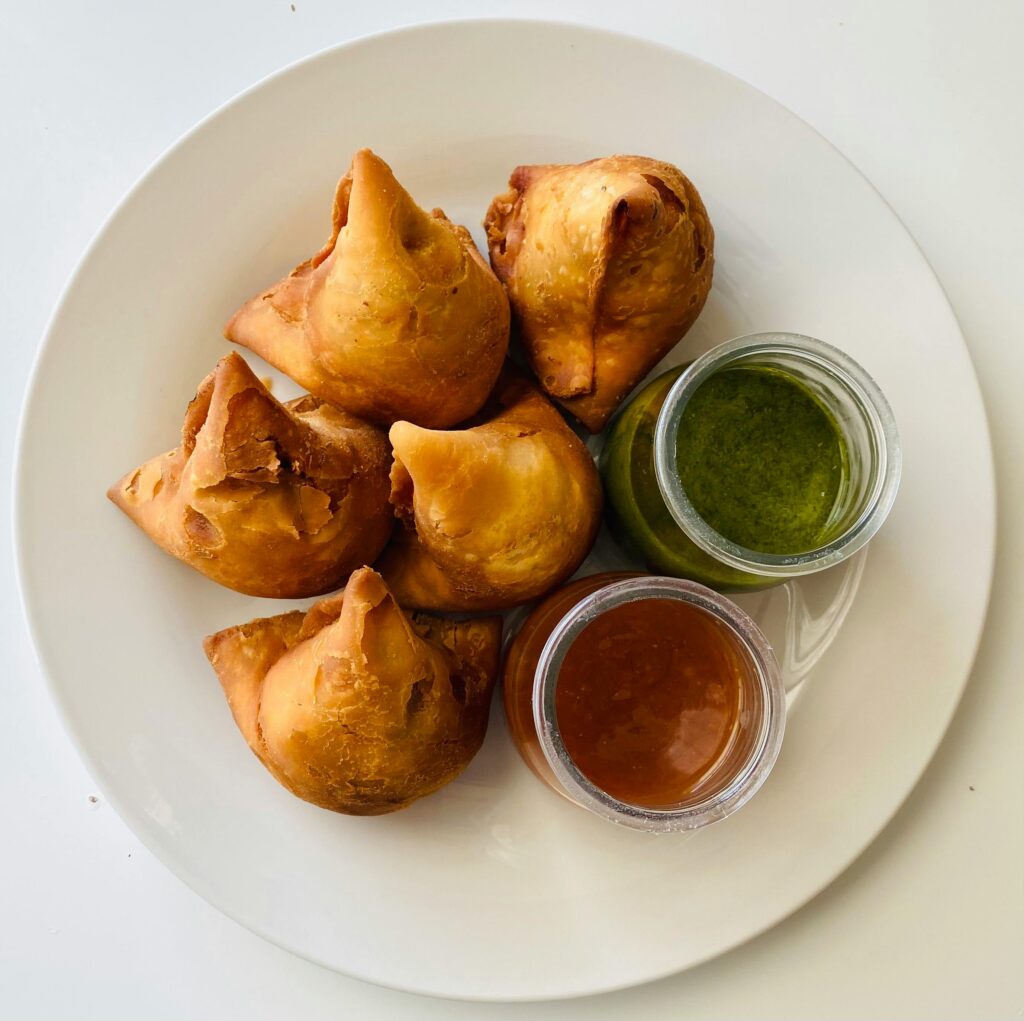 A white plate with five golden samosas, accompanied by small jars of green chutney and brown sauce, set against a plain background.
