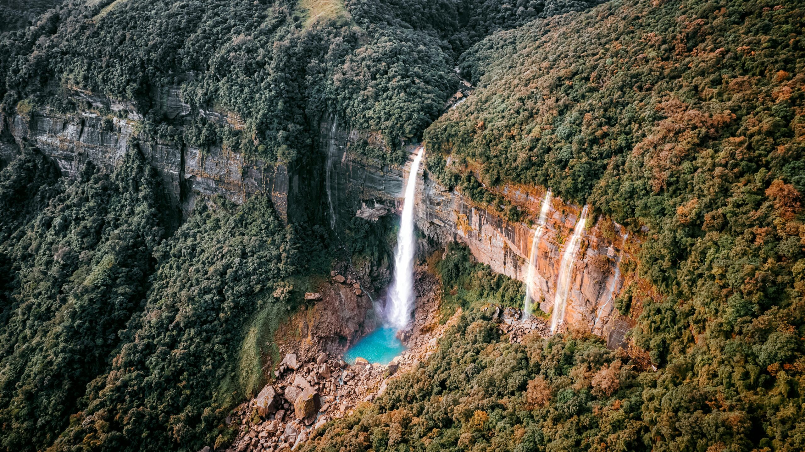 Aerial view of a stunning waterfall cascading into a turquoise pool surrounded by lush green forests and rocky cliffs in Cherrapunji, Meghalaya.